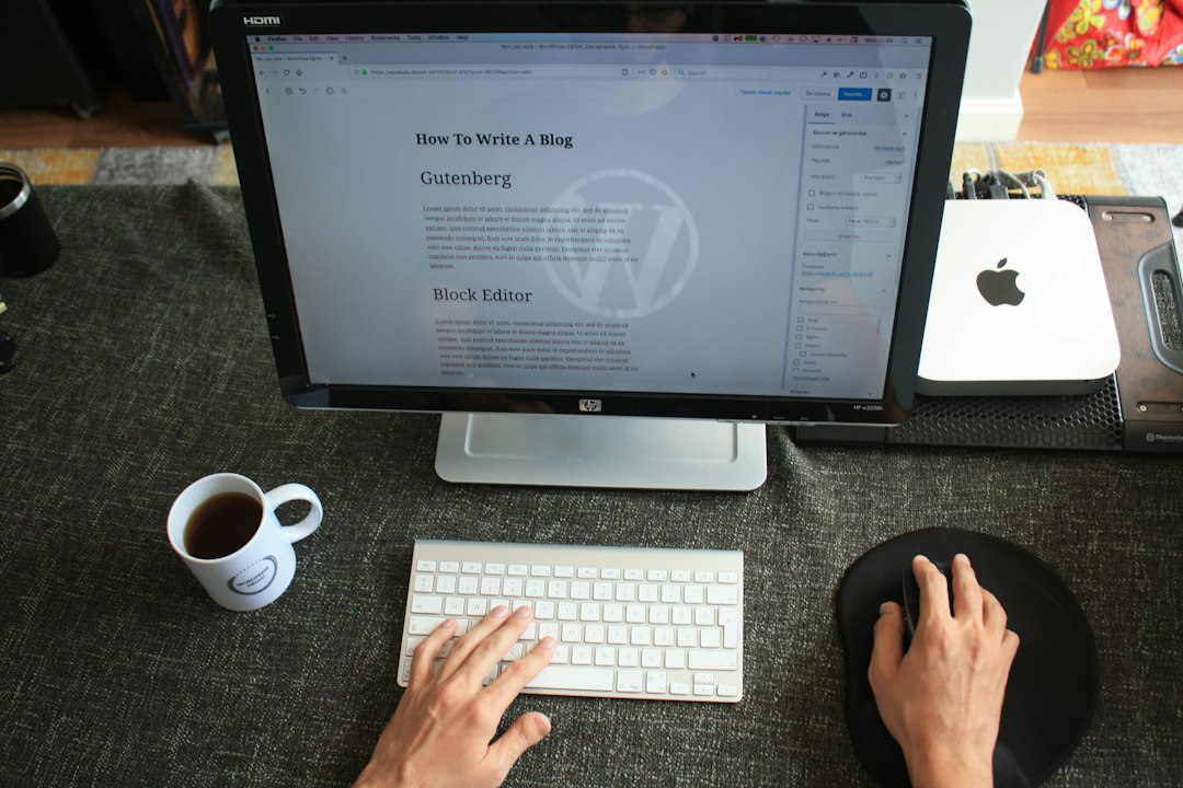 A person types on a white keyboard at a desk, editing a How to Write a Blog post about WordPress Hosting on a large monitor. A cup of coffee, mousepad, and tech accessories are neatly arranged nearby.